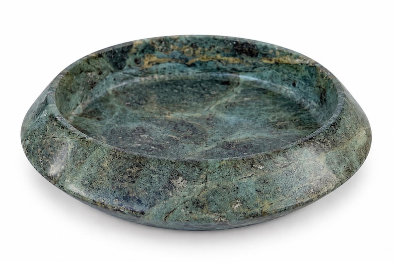 May include: A circular, shallow bowl crafted from dark green stone with natural veining and mottled patterns. The object features a wide, sloped rim and a smooth, polished surface, showcasing deep forest green tones with subtle black and grey inclusions.