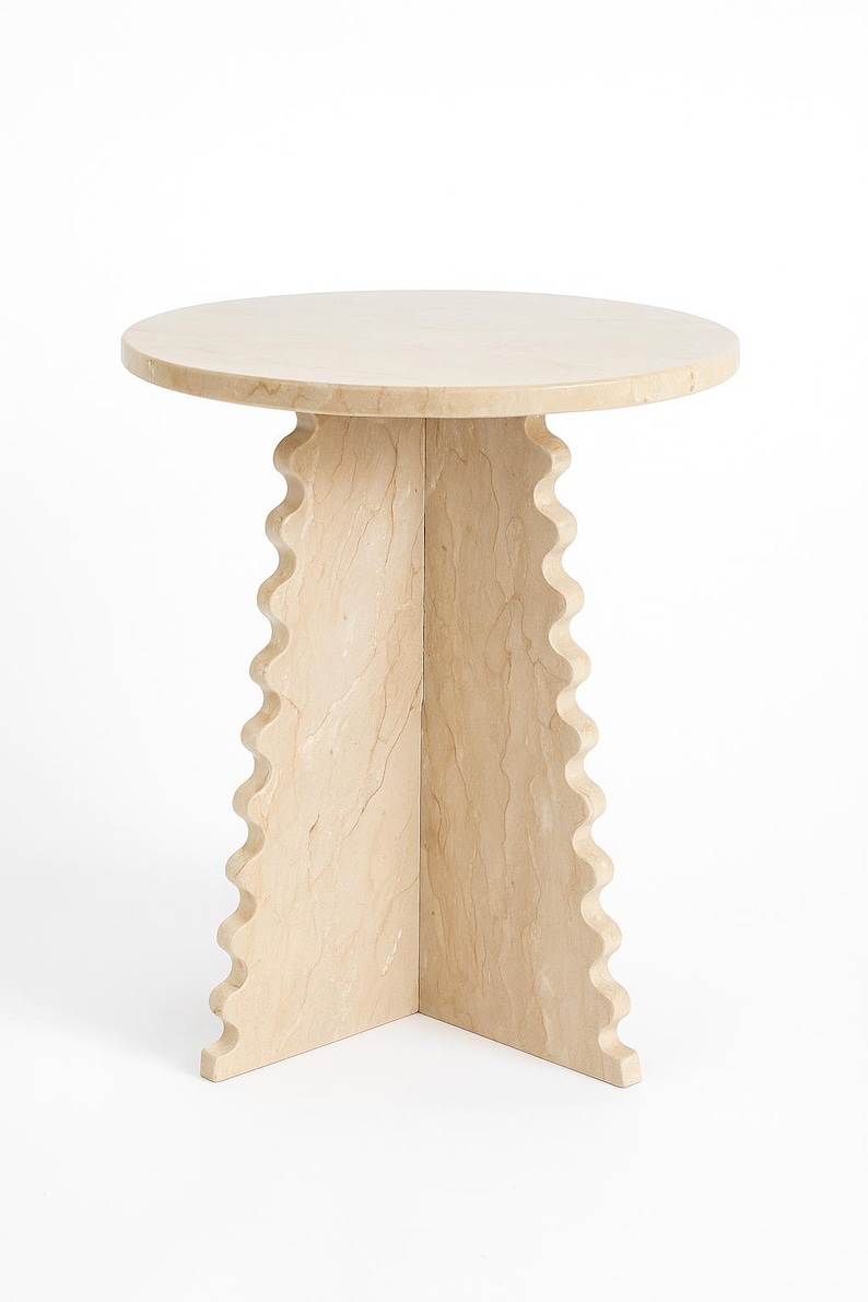 Pode incluir: Uma mesa lateral redonda bege com um design &uacute;nico. O tampo da mesa &eacute; liso e a base apresenta uma borda ondulada e recortada. A mesa &eacute; feita de um material de pedra de cor clara e tem aproximadamente 50 cm de di&acirc;metro.
