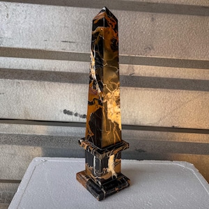 May include: A tall, four-sided obelisk sculpture crafted from polished stone featuring a striking marbled pattern of deep black, warm amber, and creamy white veins. The structure stands on a square base with a tiered pedestal design.