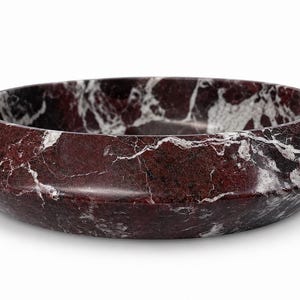 May include: A circular bowl crafted from deep burgundy marble with prominent white veining. The bowl features a wide, angled rim that slopes inward toward the center, showcasing a polished, smooth surface with natural stone patterns throughout.