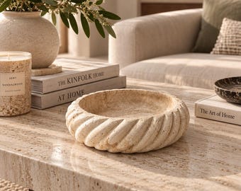 Puede incluir: Un cuenco redondo de travertino beige con un borde texturizado en forma de cuerda espiral. La superficie de piedra muestra patrones porosos naturales y variaciones de color, con un cuenco interior poco profundo y liso y una base gruesa.