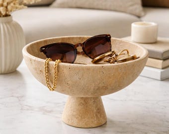 May include: A beige, pedestal bowl holds sunglasses, a gold chain, and other accessories. The bowl is made of a textured material and sits on a marble-topped table. A sofa and decorative items are in the background.