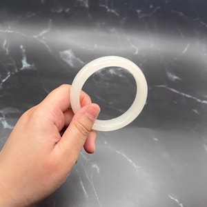 Puede incluir: Un brazalete blanco y circular se sostiene en una mano sobre un fondo oscuro. El brazalete es liso y opaco, con un color uniforme. El fondo tiene una apariencia marmolada.