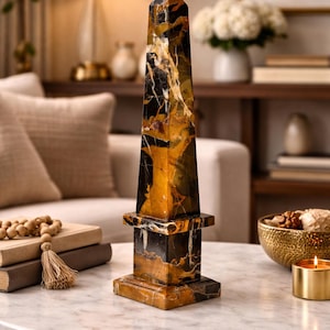 May include: A decorative obelisk with a square base and a pointed top. The obelisk is made of marble with a striking pattern of black, gold, and brown veining. It stands on a marble-topped table.