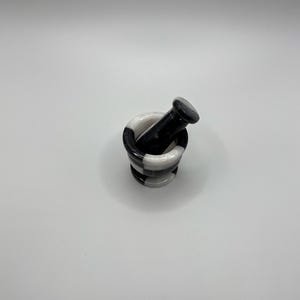 May include: A small mortar and pestle set. The mortar is cylindrical with a black and white checkered pattern. The pestle is solid black. The set is made of stone and is designed for grinding herbs and spices.