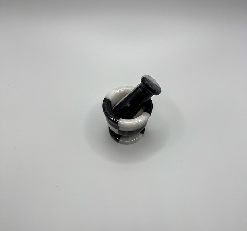 May include: A small mortar and pestle set. The mortar is cylindrical with a black and white checkered pattern. The pestle is solid black. The set is made of stone and is designed for grinding herbs and spices.