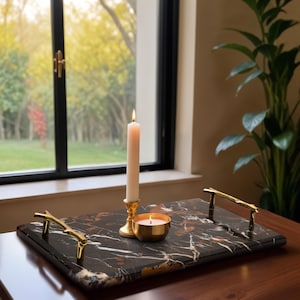 May include: A rectangular black marble tray with gold handles, holding a lit candle in a gold holder and a small gold candle. The tray is on a wooden table near a window with a view of trees.