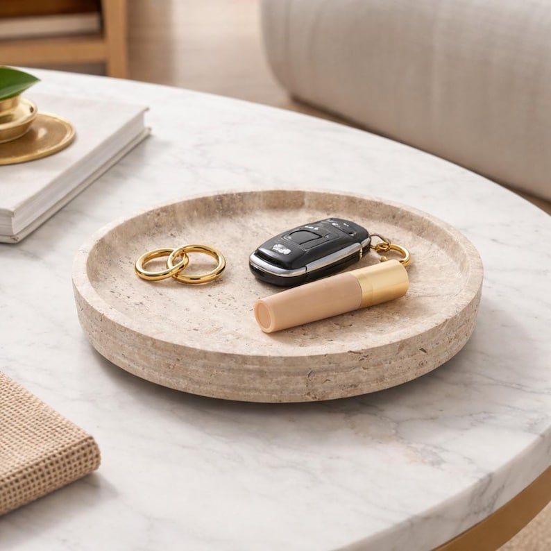 Puede incluir: Una bandeja circular de piedra beige porosa con un borde elevado. Dentro de la bandeja hay dos anillos de metal dorado, un mando de coche negro con un llavero dorado y un tubo cil&iacute;ndrico con cuerpo beige y una tapa de color dorado.
