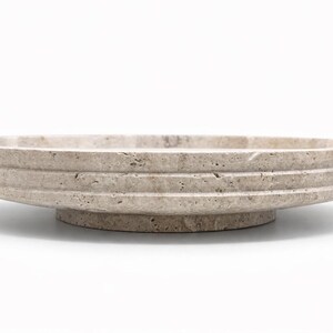 Puede incluir: Un cuenco circular de piedra travertino beige con una textura natural porosa y un dise&ntilde;o exterior escalonado. El cuenco se asienta sobre una base circular s&oacute;lida y elevada, mostrando las variaciones org&aacute;nicas de la piedra natural.