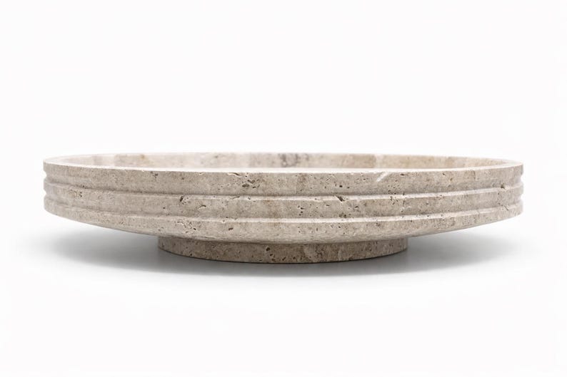 Puede incluir: Un cuenco circular de piedra travertino beige con una textura natural porosa y un dise&ntilde;o exterior escalonado. El cuenco se asienta sobre una base circular s&oacute;lida y elevada, mostrando las variaciones org&aacute;nicas de la piedra natural.