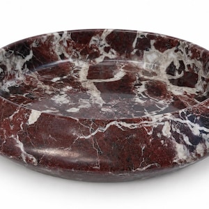 May include: A circular bowl crafted from deep red marble featuring prominent white veining throughout the stone surface. The bowl has a wide, smooth, rounded rim and a shallow interior basin, showcasing the natural polished texture of the material.