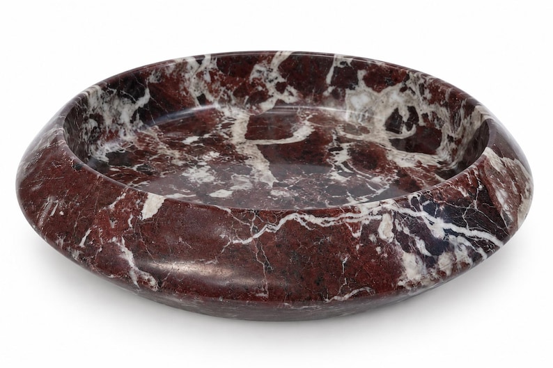 May include: A circular bowl crafted from deep red marble featuring prominent white veining throughout the stone surface. The bowl has a wide, smooth, rounded rim and a shallow interior basin, showcasing the natural polished texture of the material.