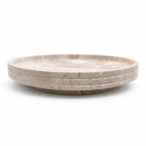 Puede incluir: Una bandeja circular de piedra travertino beige con una textura natural porosa. La bandeja presenta un borde elevado con dos l&iacute;neas horizontales ranuradas a lo largo del borde exterior y una base ligeramente hundida de perfil bajo.