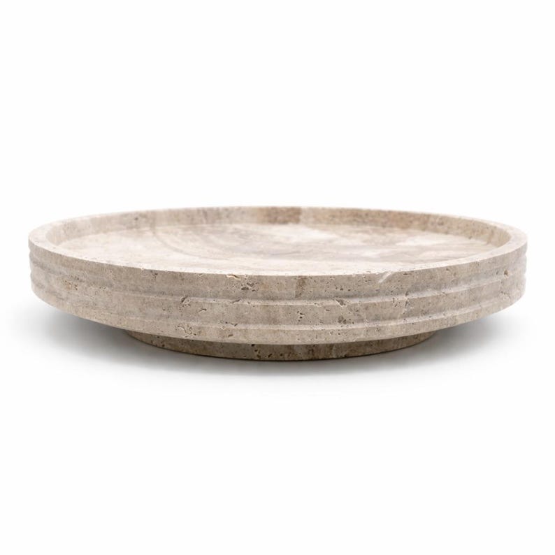 Puede incluir: Una bandeja circular de piedra travertino beige con una textura natural porosa. La bandeja presenta un borde elevado con dos l&iacute;neas horizontales ranuradas a lo largo del borde exterior y una base ligeramente hundida de perfil bajo.