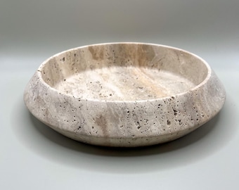 May include: A circular bowl crafted from beige travertine stone, featuring a porous, textured surface with natural veining and a wide, angled rim. The bowl has a shallow interior and a solid, heavy appearance with neutral earth tones throughout.