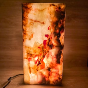 May include: A rectangular onyx lamp with a warm, glowing interior. The lamp's exterior features a blend of orange, red, and cream colors, creating a marbled effect. A black power cord extends from the base. The lamp is lit, showcasing the stone's natural patterns.
