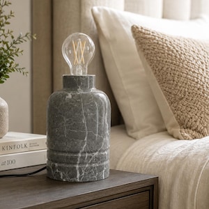 May include: A cylindrical table lamp base crafted from grey marble with white veining, featuring a smooth finish and three horizontal grooves near the bottom. The lamp is topped with a clear glass Edison-style light bulb with visible filaments.