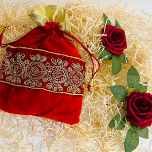 May include: A red velvet drawstring bag with gold trim and a decorative embroidered border. The bag is surrounded by shredded paper and two red roses.