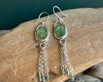 May include: Silver earrings with green gemstone drops and dangling chains. The earrings have a simple oval design and are accented with smaller green gemstones.