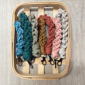 May include: A wooden basket holds several macrame wrist straps in various colors, including blue, green, tan, and pink. Each strap has a black clasp. The straps are arranged in a row, showcasing their braided design.