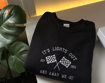 Peut inclure: Un sweat-shirt noir à col rond avec une broderie blanche représentant deux drapeaux de course à damier croisés. Le texte indique IT'S LIGHTS OUT, EST 1950, AND AWAY WE GO. Le motif est centré sur la poitrine du vêtement plié sur une surface blanche.