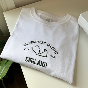 Pode incluir: Uma camisola branca de gola redonda com bordado preto no peito. O design inclui o texto "SILVERSTONE CIRCUIT", "EST 1948" e "ENGLAND" em torno de um contorno preto do mapa do circuito de corridas de Silverstone.