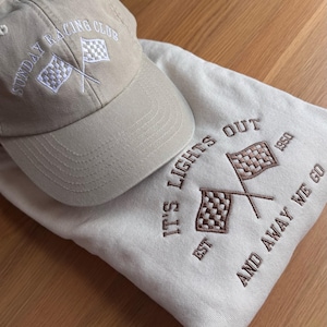 May include: A sand sweatshirt with the text "IT'S LIGHTS OUT EST 1950 AND AWAY WE GO EST 1950 AND AWAY WE GO" embroidered in light brown. The sweatshirt features two crossed checkered flags and a sand cap with the text “Sunday Racing Club” ebroidered in white.