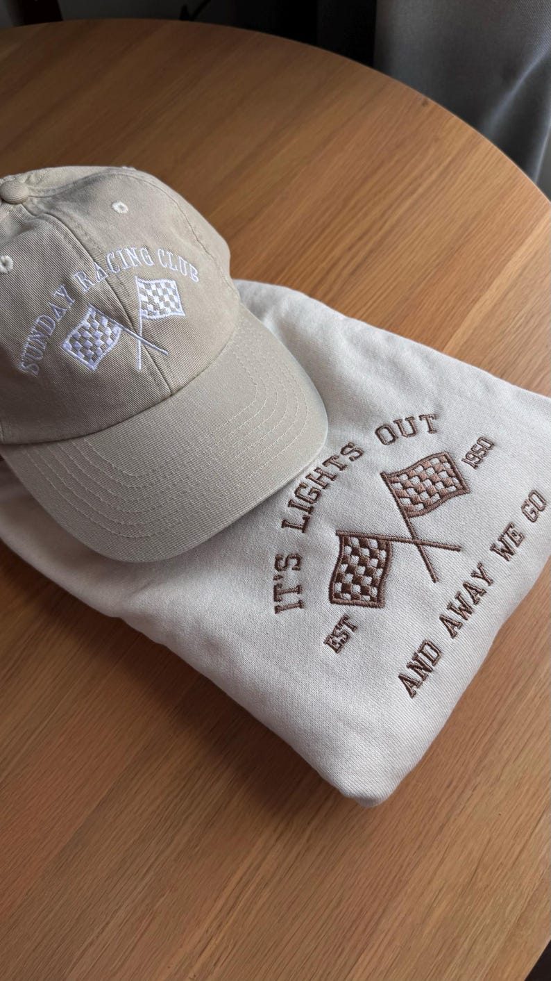 May include: A sand sweatshirt with the text "IT'S LIGHTS OUT EST 1950 AND AWAY WE GO EST 1950 AND AWAY WE GO" embroidered in light brown. The sweatshirt features two crossed checkered flags and a sand cap with the text “Sunday Racing Club” ebroidered in white.