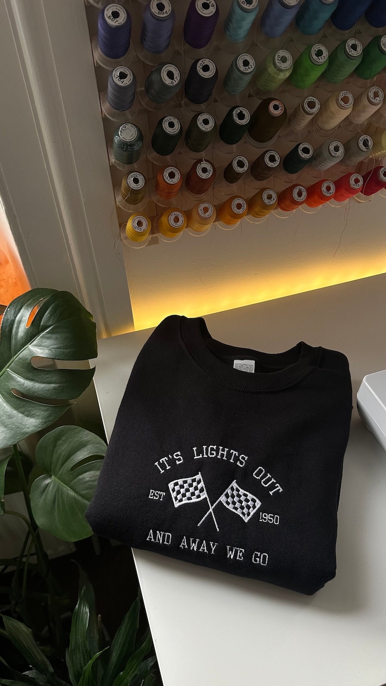 K&ouml;nnte beinhalten: Schwarzer Pullover mit wei&szlig; gesticktem Text, der "It's Lights Out Est. 1950 And Away We Go" lautet, mit einem karierten Flaggen-Motiv.