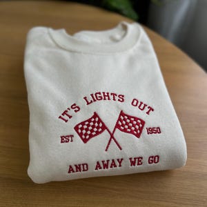 K&ouml;nnte beinhalten: Ein wei&szlig;er Pullover mit rotem gesticktem Text, der "It's Lights Out Est 1950 And Away We Go" lautet. Der Text ist von zwei roten karierten Flaggen umgeben.
