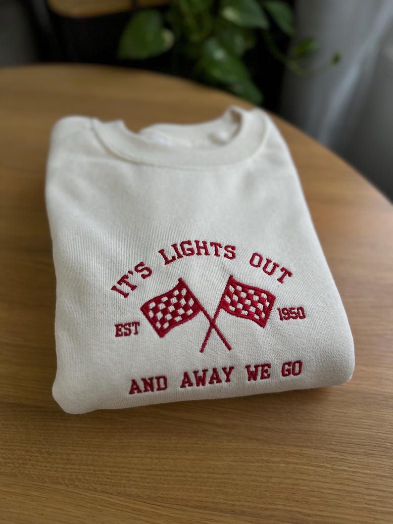 K&ouml;nnte beinhalten: Ein wei&szlig;er Pullover mit rotem gesticktem Text, der "It's Lights Out Est 1950 And Away We Go" lautet. Der Text ist von zwei roten karierten Flaggen umgeben.
