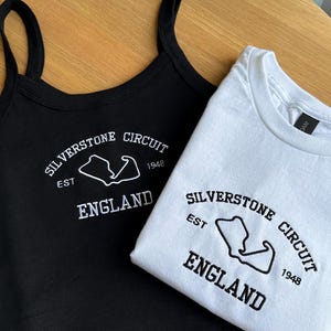 Pode incluir: Uma regata preta e uma t-shirt branca, ambas com uma estampa gr&aacute;fica que diz Silverstone Circuit, Est 1948, England, juntamente com o contorno do circuito. A regata tem al&ccedil;as finas e a t-shirt tem um decote redondo.