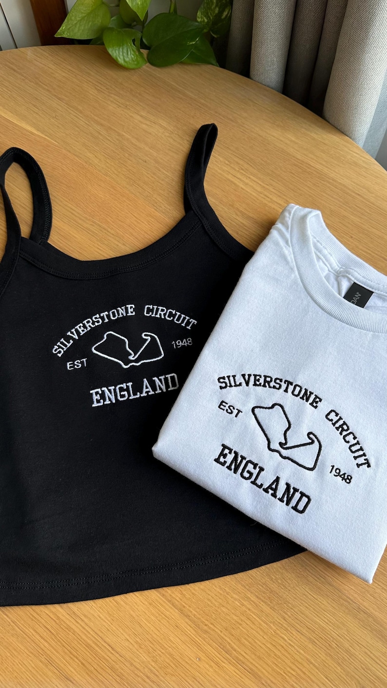 Pode incluir: Uma regata preta e uma t-shirt branca, ambas com uma estampa gr&aacute;fica que diz Silverstone Circuit, Est 1948, England, juntamente com o contorno do circuito. A regata tem al&ccedil;as finas e a t-shirt tem um decote redondo.