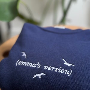 May include: A navy blue sweatshirt with white embroidered text that reads "(emma's version)" and three white embroidered birds.