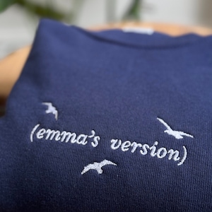 May include: A navy blue sweatshirt with white embroidered text that reads "(emma's version)" and white embroidered birds.