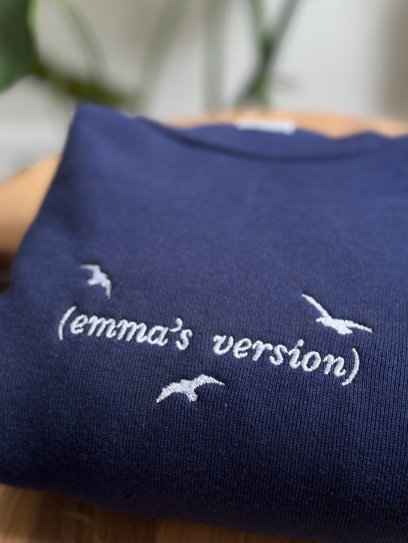 May include: A navy blue sweatshirt with white embroidered text that reads "(emma's version)" and white embroidered birds.