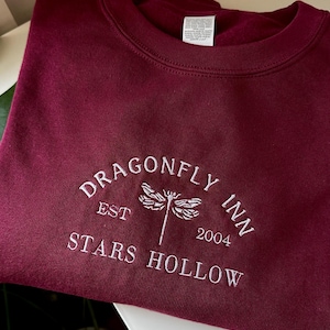 May include: Burgundy crewneck sweatshirt with white embroidered text and a dragonfly design. The text reads "DRAGONFLY INN EST 2004 STARS HOLLOW". A cozy, casual top for fans of the fictional town.