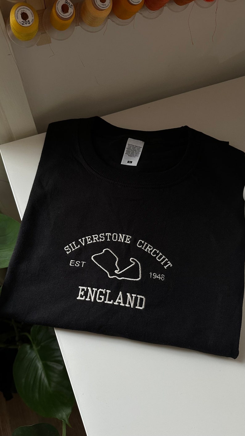 Pode incluir: Uma camisola preta de gola redonda com bordado branco no peito. O design inclui o texto "SILVERSTONE CIRCUIT", "EST 1948" e "ENGLAND" em torno de um contorno branco do mapa do circuito de corridas de Silverstone.