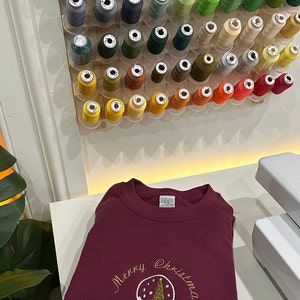 Pu&ograve; includere: Una felpa color bordeaux con ricamo dorato di una palla di neve con un albero di Natale all'interno e il testo "Merry Christmas".