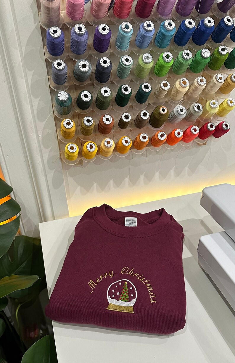 Pu&ograve; includere: Una felpa color bordeaux con ricamo dorato di una palla di neve con un albero di Natale all'interno e il testo "Merry Christmas".