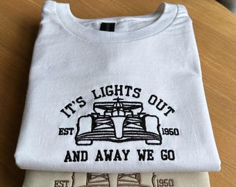 May include: A white crewneck sweatshirt with black embroidery featuring a Formula One race car, the text IT'S LIGHTS OUT AND AWAY WE GO, and EST 1950. Beneath it, a beige sweatshirt features the same design embroidered in a matching beige thread.