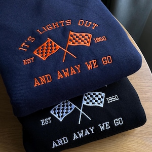 K&ouml;nnte beinhalten: Zwei Sweatshirts mit Rennthema. Das obere Sweatshirt ist marineblau mit orangefarbener Stickerei. Das untere Sweatshirt ist schwarz mit wei&szlig;er Stickerei. Beide Sweatshirts zeigen gekreuzte karierte Flaggen, den Text "It's Lights Out", "Est. 1950" und "And Away We Go".