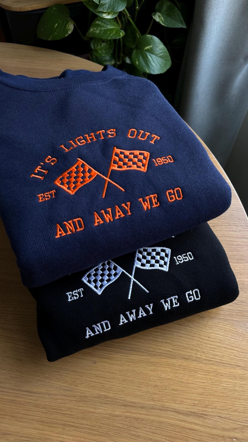 K&ouml;nnte beinhalten: Zwei Sweatshirts mit Rennthema. Das obere Sweatshirt ist marineblau mit orangefarbener Stickerei. Das untere Sweatshirt ist schwarz mit wei&szlig;er Stickerei. Beide Sweatshirts zeigen gekreuzte karierte Flaggen, den Text "It's Lights Out", "Est. 1950" und "And Away We Go".