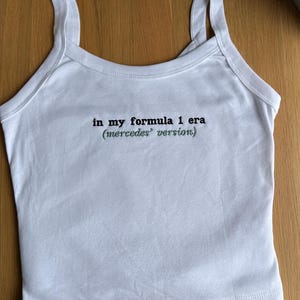 May include: A white sleeveless vest top with a scoop neckline and thick shoulder straps. The centre of the chest features black embroidered text that reads: in my formula 1 era (mercedes version). The garment is made of a solid white fabric.