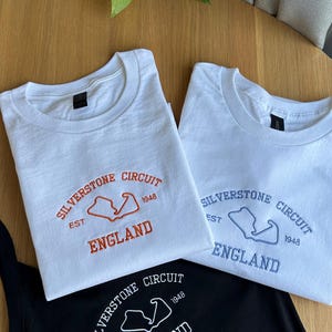 Pode incluir: Tr&ecirc;s t-shirts com texto bordado e o contorno de um circuito. Uma t-shirt branca tem bordado laranja com Silverstone Circuit, Est 1948, England. Outra t-shirt branca tem bordado azul e uma t-shirt preta tem bordado branco.