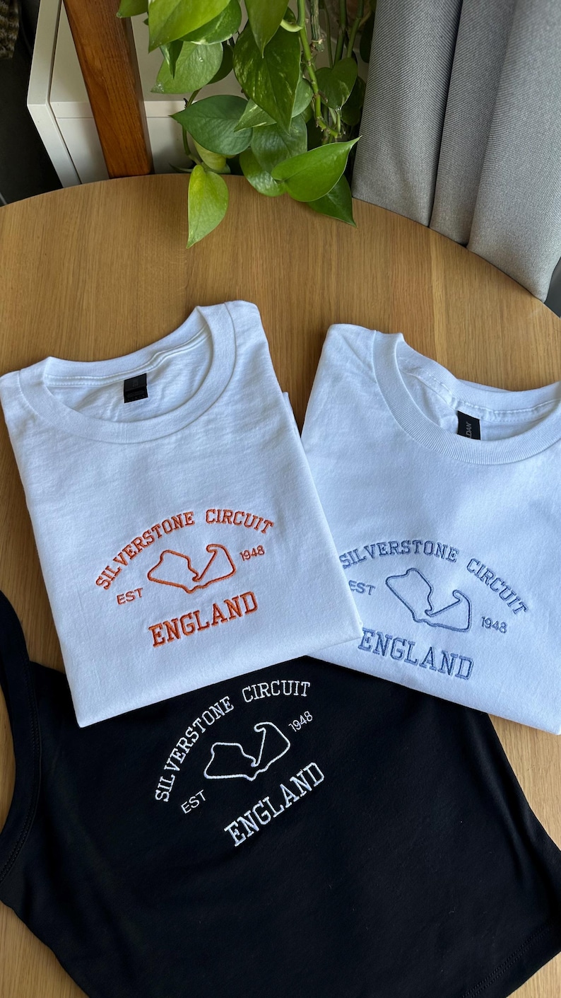 Pode incluir: Tr&ecirc;s t-shirts com texto bordado e o contorno de um circuito. Uma t-shirt branca tem bordado laranja com Silverstone Circuit, Est 1948, England. Outra t-shirt branca tem bordado azul e uma t-shirt preta tem bordado branco.
