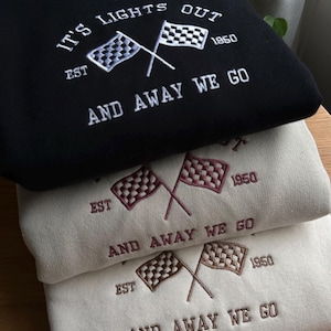 K&ouml;nnte beinhalten: Drei Sweatshirts &uuml;bereinander gestapelt. Das oberste Sweatshirt ist schwarz mit wei&szlig;er Stickerei, die "IT'S LIGHTS OUT EST 1950 AND AWAY WE GO" lautet. Das mittlere Sweatshirt ist beige mit burgunderfarbener Stickerei, die "EST 1950 AND AWAY WE GO" lautet. Das unterste Sweatshirt ist beige mit brauner Stickerei, die "EST 1950 AND AWAY WE GO" lautet.