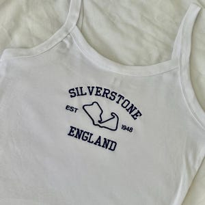 Pode incluir: Uma regata branca com texto bordado em azul escuro que diz Silverstone England, com a abrevia&ccedil;&atilde;o EST e o ano 1948 ladeando um contorno azul escuro do mapa do circuito de corrida de Silverstone centralizado no peito.