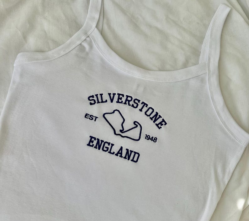 Pode incluir: Uma regata branca com texto bordado em azul escuro que diz Silverstone England, com a abrevia&ccedil;&atilde;o EST e o ano 1948 ladeando um contorno azul escuro do mapa do circuito de corrida de Silverstone centralizado no peito.