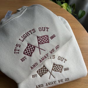 K&ouml;nnte beinhalten: Ein hellgraues Sweatshirt mit dem Text "IT'S LIGHTS OUT EST 1950 AND AWAY WE GO EST 1950 AND AWAY WE GO", der in Maroon gestickt ist. Das Sweatshirt zeigt zwei gekreuzte karierte Flaggen, eine in Maroon und eine in Braun.